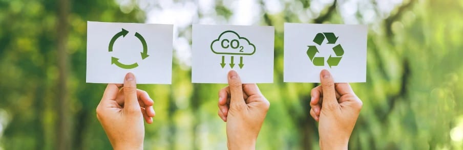 Drie handen houden kaarten vast tegen een onscherpe, groene natuurlijke achtergrond; de kaarten tonen symbolen voor recyclen, CO2-reductie en het algemene recyclinglogo, die samenwerken aan duurzaamheid en milieubewustzijn.