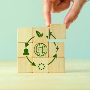 Een hand plaatst een houten blokje op een stapel van blokken, die samen een cirkel van duurzaamheidssymbolen vormen, waaronder een wereldbol, bladeren, pijlen en tandwielen, die de circulaire economie vertegenwoordigen.