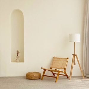 Een minimalistisch ingerichte kamer met een rotan stoel, een bijpassend poefje, een houten vloerlamp met witte kap en een nis in de muur met een vaas met droogbloemen allemaal onderdeel van circulaire economie